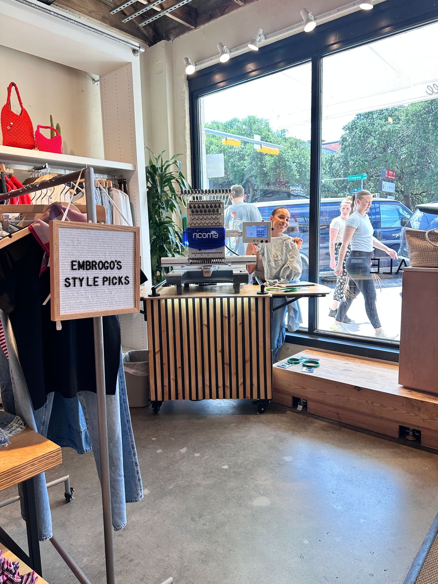 A mobile embroidery station featuring a Ricoma machine is set up inside a trendy boutique next to a large window facing the street. A smiling staff member holds up a freshly embroidered garment as pedestrians walk by outside. A sign reads “EMBROGO’S STYLE PICKS” beside a curated clothing rack with denim and fashion-forward pieces. The setup blends retail and customization, highlighting a live embroidery activation in a stylish, urban shopping environment.