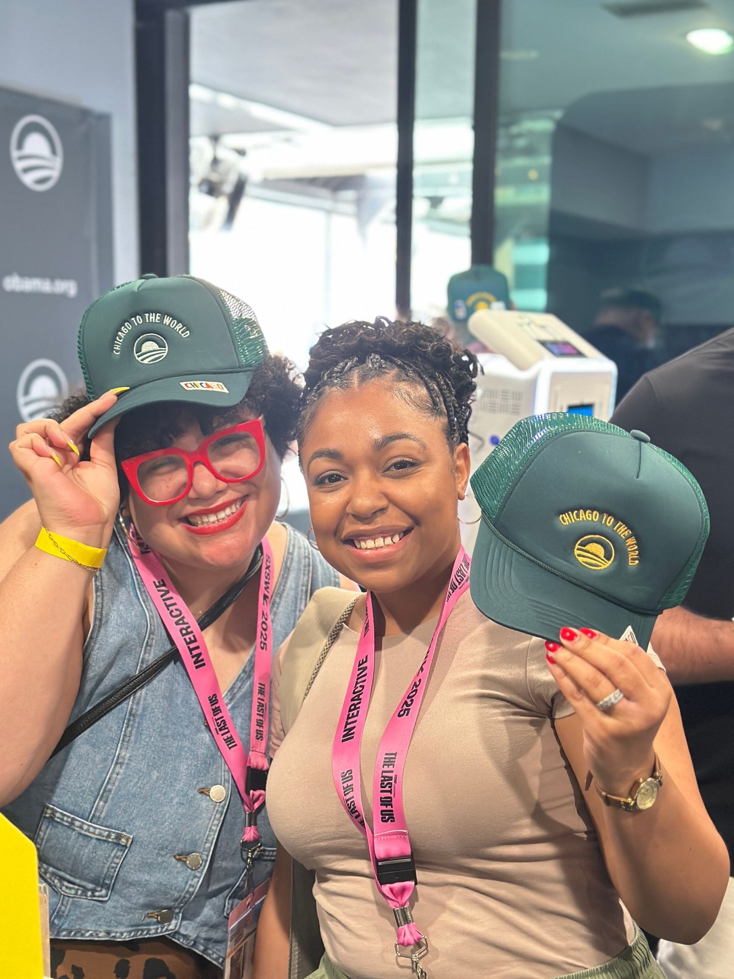  Two smiling women proudly showcase custom green trucker hats embroidered with “CHICAGO TO THE WORLD” at a live event. They wear interactive event lanyards from “The Last Sip” 2025, standing in front of a modern booth setup with embroidery equipment in the background. One woman wears bold red glasses and denim; the other holds a hat and wears a neutral-toned outfit. The atmosphere is vibrant, community-focused, and creative.